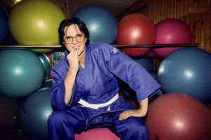 Ein Mann sitzt in einem blauen Judoanzug auf einem Gymnastikball und lächelt in die Kamera. Hinter ihm sind lauter bunte Gymnastikbälle.